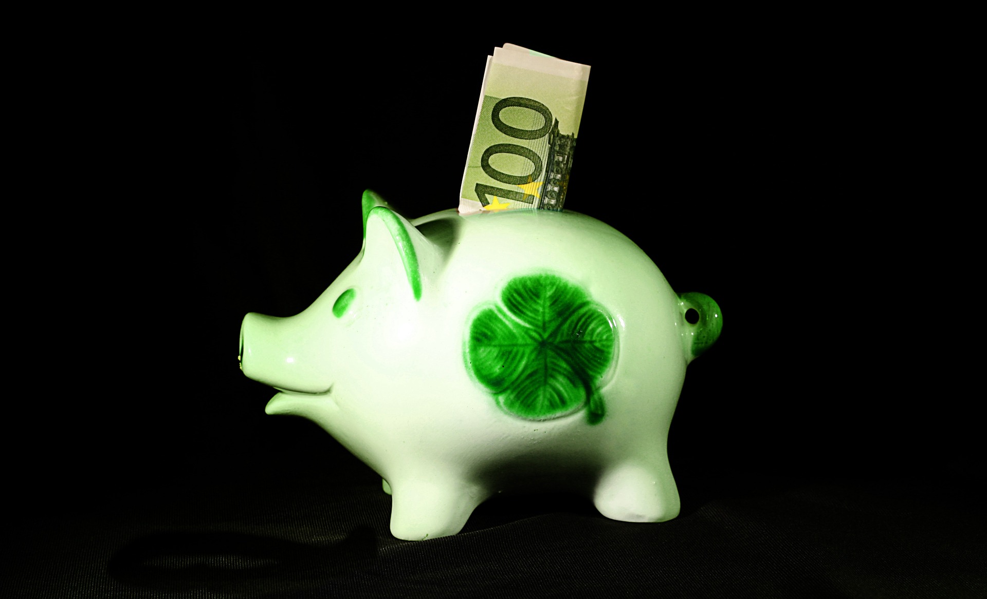 In einem weißen Sparschwein mit einem Kleeblatt befinden sich 100 Euro im Einwurf-Schlitz.